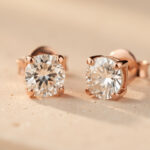 Quelles boucles d’oreilles en diamant champagne choisir selon leur qualité ? Gros plan de boucles d’oreilles puces en diamant champagne serties sur de l’or rose, présentées sur un fond beige minéral avec une lumière naturelle douce.
